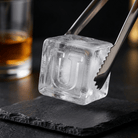 Metal tongs gripping a large clear ice cube stamped with the letter U over a dark slate coaster, with a glass of whiskey in the background.