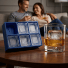 A blue silicone Letter V ice tray and a whiskey glass holding a clear ice cube stamped with the letter V on a wooden table, with a couple relaxing in the background.