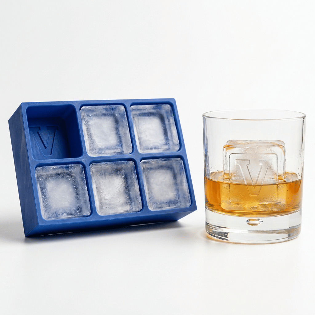 A blue silicone Letter V ice tray beside a whiskey glass containing amber liquor and a clear ice cube stamped with the letter V.