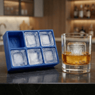A blue silicone Letter W ice tray beside a whiskey glass holding a clear ice cube stamped with the letter W, set on a dark bar counter with backlit bottles behind.