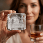 A woman holds a large clear ice cube stamped with the letter W up to the camera, with a glass of whiskey in the background.