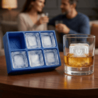 A blue silicone Letter X ice tray and a whiskey glass holding a clear ice cube stamped with the letter X on a wooden table, with a couple relaxing in the background.