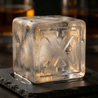 A large clear ice cube stamped with the letter X resting on a dark slate coaster, with a glass of whiskey blurred in the background.