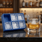 A blue silicone Letter Y ice tray beside a whiskey glass holding a clear ice cube stamped with the letter Y, set on a dark bar counter with backlit bottles behind.