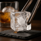 Metal tongs gripping a large clear ice cube stamped with the letter Y over a dark slate coaster, with a glass of whiskey in the background.
