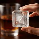 A hand holds a large clear ice cube stamped with the letter Z, with a glass of whiskey in the background.